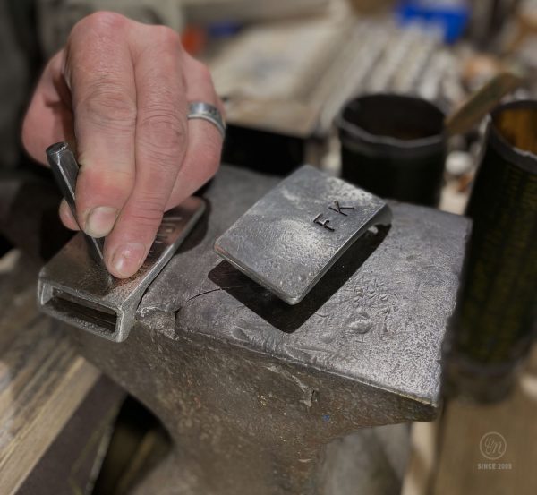 De afbeelding toont in detail een op maat gemaakte riemgesp gemaakt door Neptunsgems. De blanco riemgesp ligt op een aambeeld, je ziet een hand die een stalen reliëfstempel vasthoudt. Met deze stempels kunnen letters en cijfers in de gesp worden gegraveerd. Op de voorgrond is een riemgesp met initialen erin gestempeld.