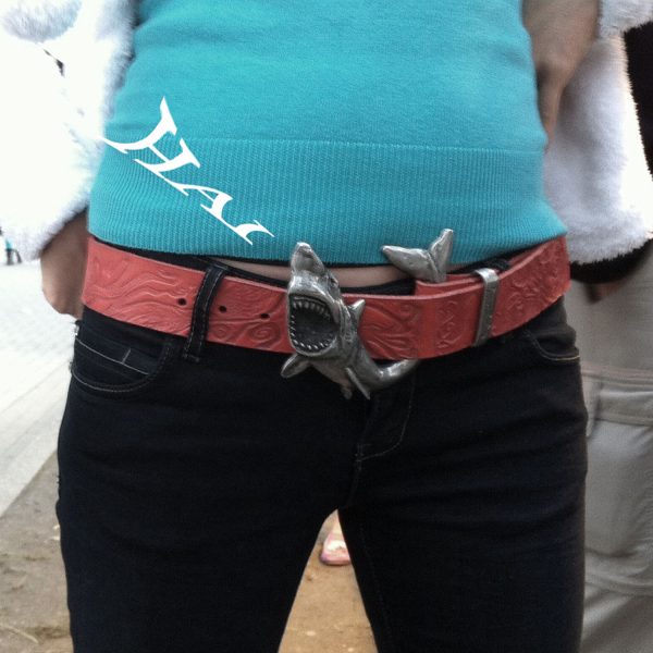 La photo montre la boucle de ceinture de requin de Neptunsschmeide sur la ceinture d'un modèle avec une ceinture en cuir rouge et un gaufrage de pieuvre. La boucle de ceinture de requin a été fabriquée à la main dans l'atelier Neptunsgemeinde à partir d'un alliage d'étain de haute qualité.