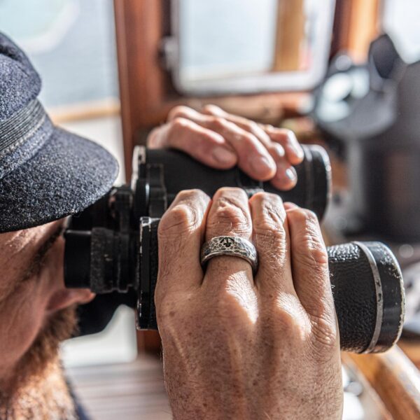 Band ring with a hammered wave surface on the finger of a captain's hand holding binoculars. Size approx. 16 x 25 mm. Sample image of Neptune jewelry.