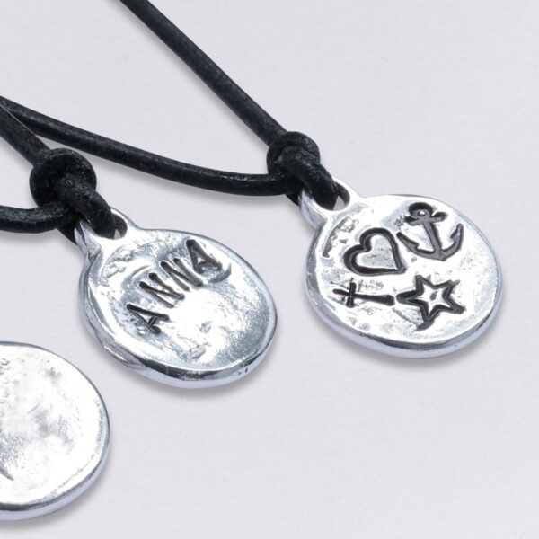 Beispiele von Wunschprägungen. Anker, Herz, Kreuz, ein Name: Anna, auf der Rückseiten von Schmuck Anhängern am Lederband von Neptunsgeschmeide.