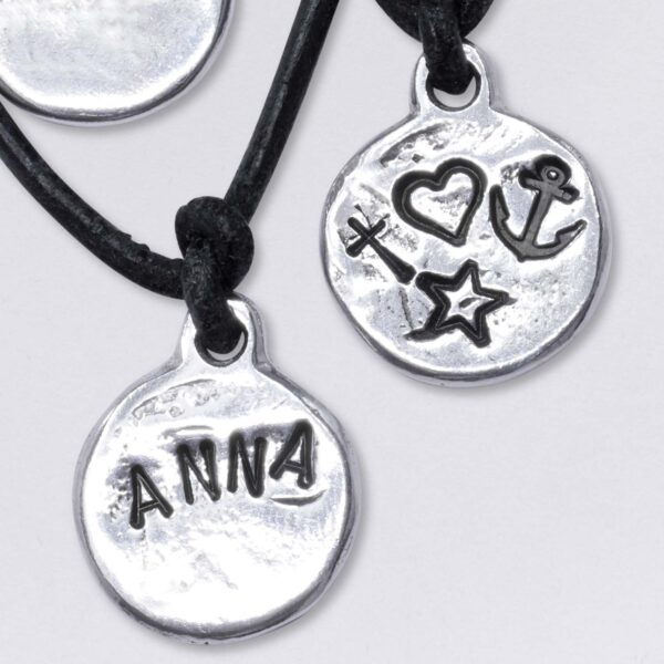 Beispiele von Wunschprägungen. Anker, Herz, Kreuz, ein Name: Anna, auf der Rückseiten von Schmuck Anhängern am Lederband von Neptunsgeschmeide in Aufsicht.