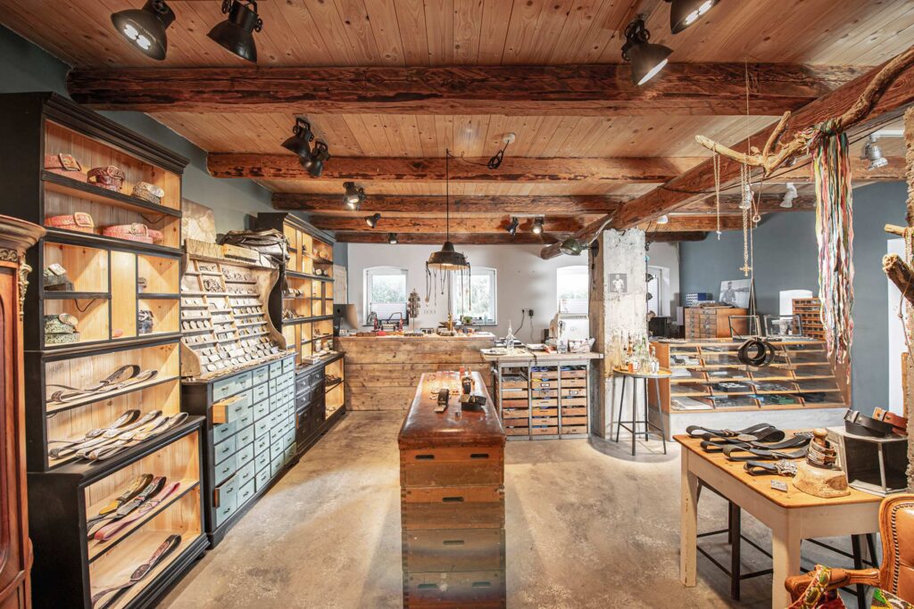 Neptunsgeschmeide Showroom. Ladenbereich, Aufnahme des Ladenbereiches ohne Kunden. Regale mit Motiv Gürtelschließen, Lederriemen und Schmuck in der alten Scheune in Stampe bei Kiel..