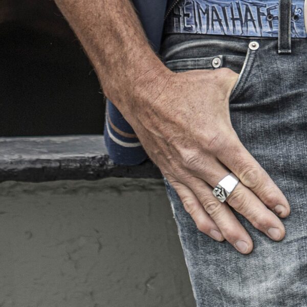 Siegelring mit Anker Motiv, geschwärzt. Größe ca. 16 x 25 mm, von Neptunsgeschmeide. Werbebild Ring an Hand, männliche Person in Jeans. Detailansicht Hand an Hose.