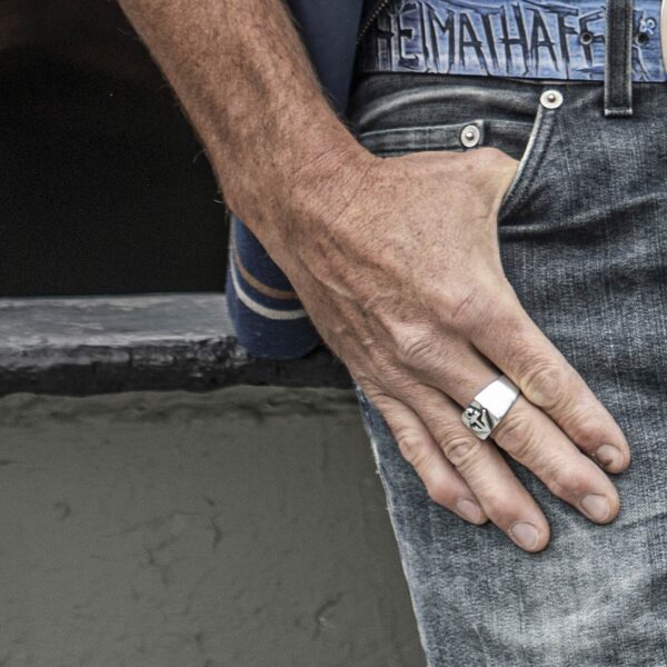 Anello con sigillo con motivo ad ancora, brunito. Dimensioni circa 16 x 25 mm, realizzato con gioielli Nettuno. Immagine promozionale dell'anello a portata di mano, persona di sesso maschile in jeans. Vista dettagliata della mano sui pantaloni.