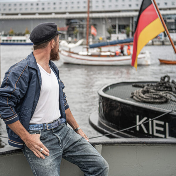 Ceinture bleue scintillante Heimathafen Kiel avec une simple boucle : pont supérieur d'environ 6 x 5 cm. L'image montre la ceinture sur le pantalon d'un marin à bord..