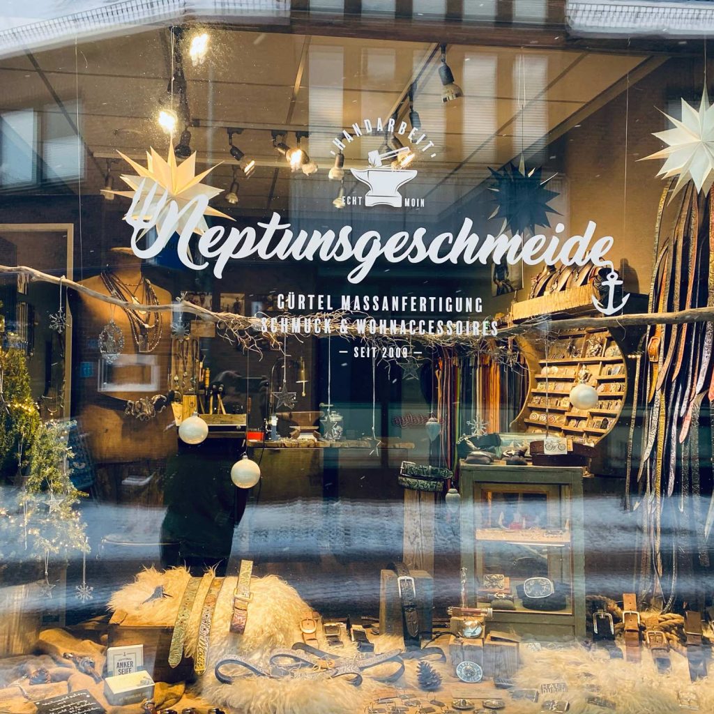 Bild zeigt das Schaufenster des kleinen Ladengeschäftes von Neptunsgeschmeide in Eckernförde. Angebracht ist eine Fenster Beklebung mit dem Neptunsgeschmeide Logo und Gürtel Maßanfertigung, Schmuck und Wohnaccessoires. Handarbeit seit 2009.