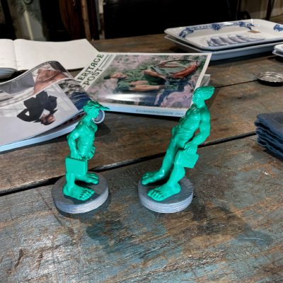 Zwei 10 cm große Miniaturen der "Reisenden Riesen im Wind" auf dem Westerländer Bahnhofsvorplatz auf Sylt stehen auf einem Tisch. Dahinter liegt eine Zeitschrift um den Größenvergleich besser darstellen zu können. Die Skulpturen wurden vom Bildhauer Martin Wolke hergestellt und sind im Neptunsgeschmeide Online Shop erhältlich.