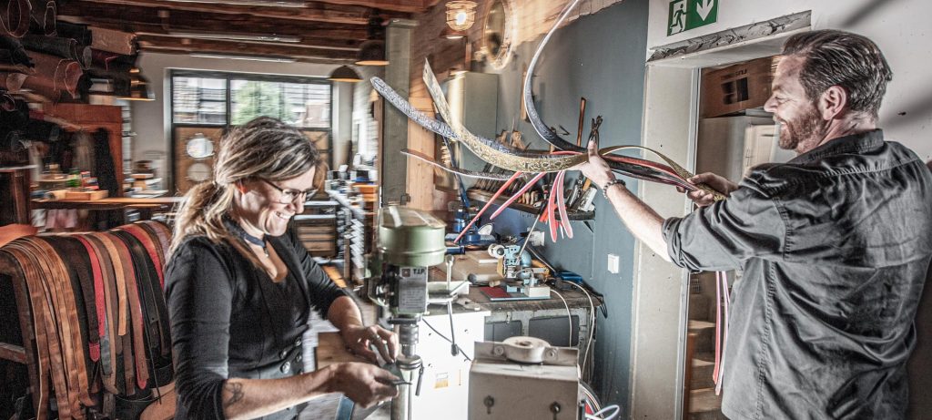 Blick in die Ledergürtel Manufaktur, Werkstatt von Neptunsgeschmeide. Gezeigt wird ein Arbeitsschritt der die Gürtelkanten poliert. Ein Bündel Gürtel wird effektvoll durch die Luft gehoben.