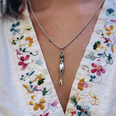 Ein Meerjungfrau-Anhänger von Neptunsgeschmeide hängt an einer Silberkette am Dekolletee einer jungen Frau mit Bluse und Blumenkragen.