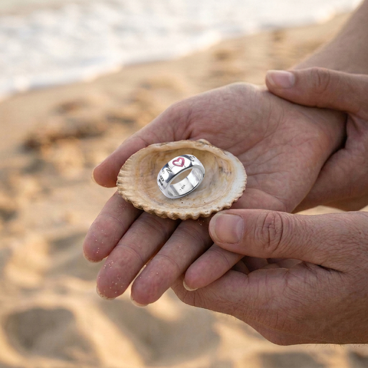 Herz Koordinaten Bandring von Neptunsgeschmeide mit polierter Oberfläche und Herzsymbol mit pinker Gießharzfüllung. Liegt auf einer Muschel, die von einem Paar in den Händen gehalten wird.